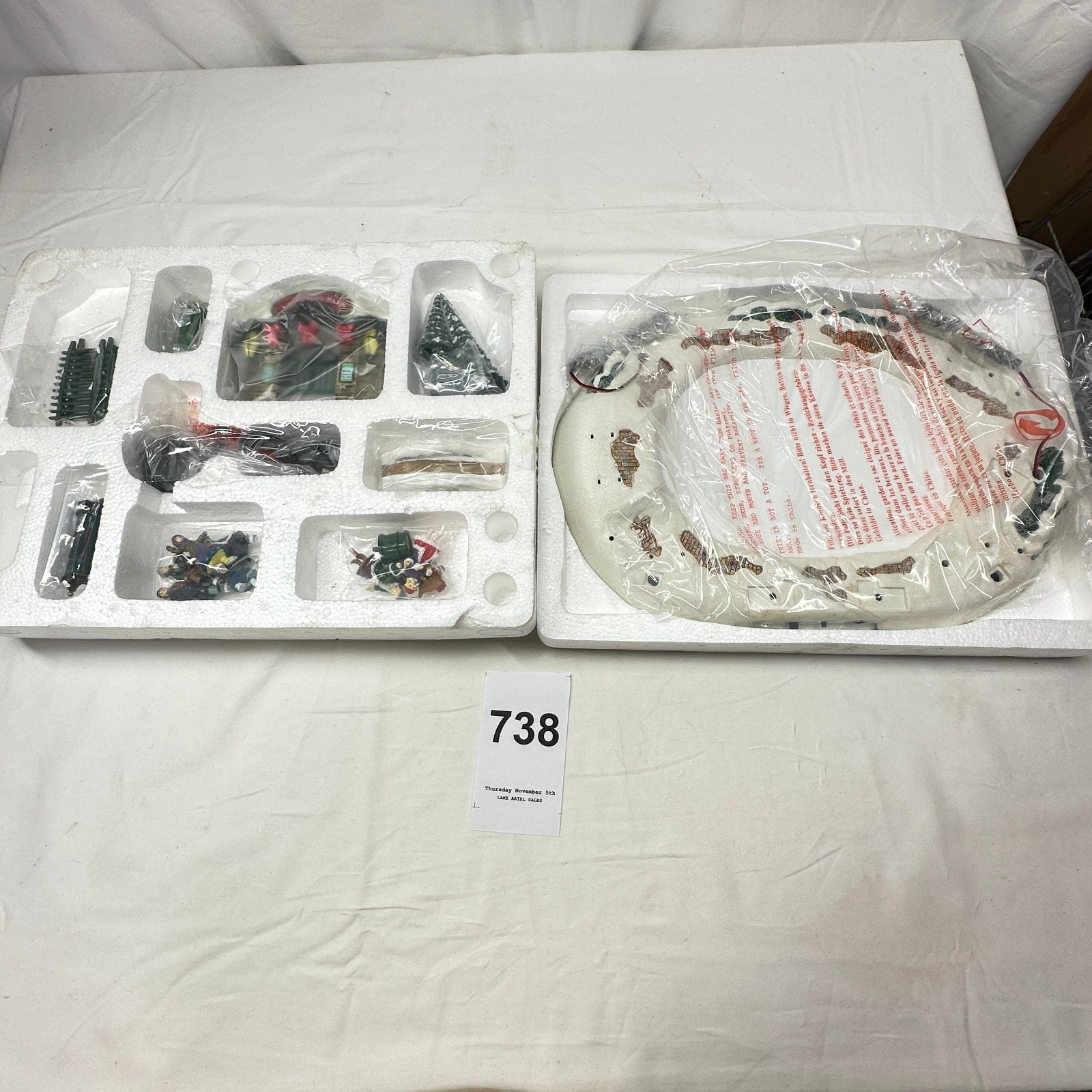 Lot 738: Christmas village accessory set, skaters figurines, lamp posts, fence, Christmas tree and street sign pieces, Department 56 style porcelain lit building, unbuilt in box