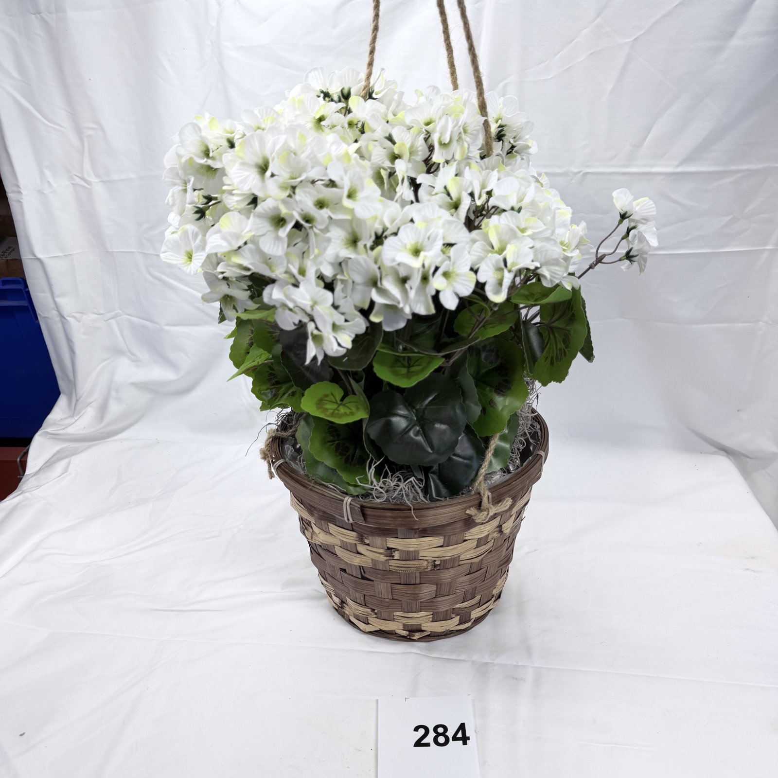 Lot 284: Artificial white geranium hanging basket arrangement in woven wicker pot with rope handles